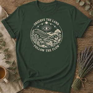 Może przedstawiać: Ciemnozielony t-shirt z krótkim rękawem z kremową grafiką w kształcie koła. Wzór przedstawia krajobraz z rzeką, wzgórzami i drzewami pod cyklem faz księżyca i symbolem oka, z napisem OBSERVE THE LAND FOLLOW THE FLOW.