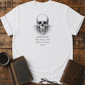 Può includere: Una t-shirt bianca a maniche corte con un'illustrazione a inchiostro nero di un teschio umano che si riflette sull'acqua. Sotto il disegno, il testo recita: REMEMBER YOU WILL DIE UNTIL THEN LIVE. La maglietta presenta un girocollo classico.