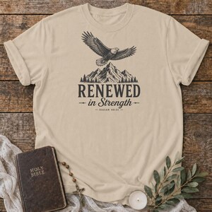 Könnte beinhalten: Ein beiges T-Shirt mit kurzen Ärmeln und einer schwarzen Grafik eines Adlers, der über ein Gebirge fliegt. Unter den Bergen steht der Text RENEWED in Strength und Isaiah 40:31 in einer Serifenschrift mittig auf der Brust.