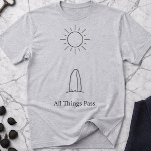 Może przedstawiać: Jasnoszara koszulka z czarną grafiką słońca i formacji skalnej. Tekst "All Things Pass" jest wydrukowany poniżej. Koszulka jest wykonana z miękkiego materiału.