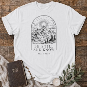 Puede incluir: Camiseta blanca con un diseño de montaña y sol en blanco y negro. El diseño está enmarcado por un arco e incluye el texto "BE STILL AND KNOW" y "PSALM 46:10".