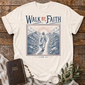 May include: Cream-colored t-shirt featuring a graphic of a person walking on a path towards a sunrise, with the words "WALK BY FAITH" and "2 COR. 5:7". The design is framed in a blue and red border.