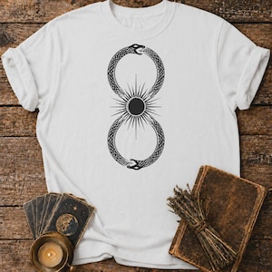 May include: A white short-sleeved t-shirt featuring a black graphic print of two snakes forming an infinity symbol shape. In the centre of the infinity loop, there is a solid black circle surrounded by radiating thin lines resembling a sunburst.