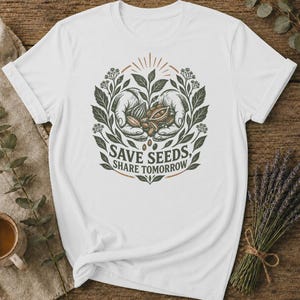 May include: A white t-shirt featuring a central graphic of two hands cupping seeds, surrounded by botanical illustrations of leaves and flowers. The text "SAVE SEEDS, SHARE TOMORROW" is printed in a dark font below the hands within the circular design.