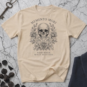 Puede incluir: Camiseta beige con una ilustración detallada de una calavera rodeada de rosas y follaje. El texto "MEMENTO MORI" se arquea sobre la calavera, con "REMEMBER WHAT MATTERS" debajo. El diseño es en tinta negra.