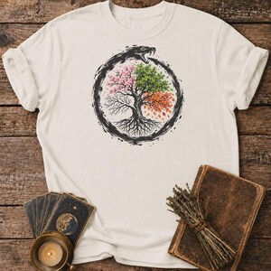 以下が含まれることがあります： クリーム色の半袖Tシャツ。4つの季節に分かれた木の円形グラフィックが描かれており、ウロボロスの形をした黒い蛇がそれを取り囲んでいる。木にはピンクの花、緑の葉、オレンジ色の秋の紅葉、そして葉のない枝が表現されている。