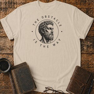 May include: A beige short-sleeved t-shirt featuring a black circular graphic on the chest. The graphic depicts a profile illustration of a bearded man with curly hair, surrounded by the text THE OBSTACLE IS THE WAY in a circular layout.
