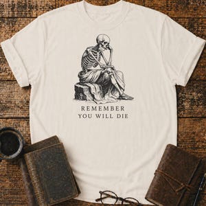 Può includere: Una t-shirt a maniche corte color crema con un'illustrazione a inchiostro nero di uno scheletro seduto su una roccia in posa pensierosa. Sotto il disegno, il testo REMEMBER YOU WILL DIE è stampato in un carattere serif chiaro e centrato.