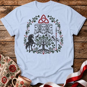 May include: Light blue t-shirt featuring a detailed graphic design. The design includes three bearded faces, a red Celtic knot, a black horse, a tree, mountains, and a spear, all within a green and red wreath. The shirt is made of a soft material.