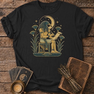 May include: Black t-shirt with an Egyptian deity illustration writing on a scroll. The design features a crescent moon, stars, and lotus flowers in gold, blue, and green. The shirt is on a wooden surface with tarot cards and a candle.