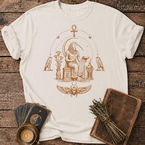 Può includere: Una t-shirt color crema con una stampa grafica marrone della divinità egizia Thot seduta su un trono, che tiene in mano un rotolo. Il design include un ankh, due falchi, fiori di loto e uno scarabeo alato centrato sotto la figura.