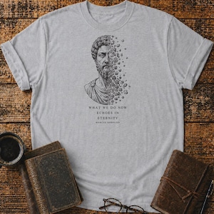 Pode incluir: Uma t-shirt cinzenta clara de manga curta com uma ilustração a tinta preta de Marco Aurélio, com o lado direito do rosto a dissolver-se em fragmentos de pedra. Abaixo do gráfico, o texto diz: WHAT WE DO NOW ECHOES IN ETERNITY MARCUS AURELIUS.