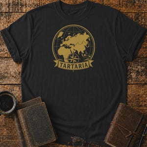 Op de afbeelding: Een zwart T-shirt met korte mouwen voorzien van een goudkleurige afbeelding van een wereldbol met een kompasroos en een banner eronder met de tekst TARTARIA in hoofdletters. Het ontwerp is gecentreerd op de borst van het kledingstuk.