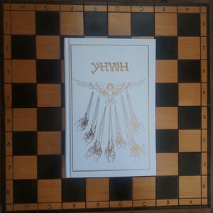 May include: A white book with gold lettering and a detailed illustration of a bird and hands, resting on a wooden chessboard with black and tan squares. The book's title is "YHWH".
