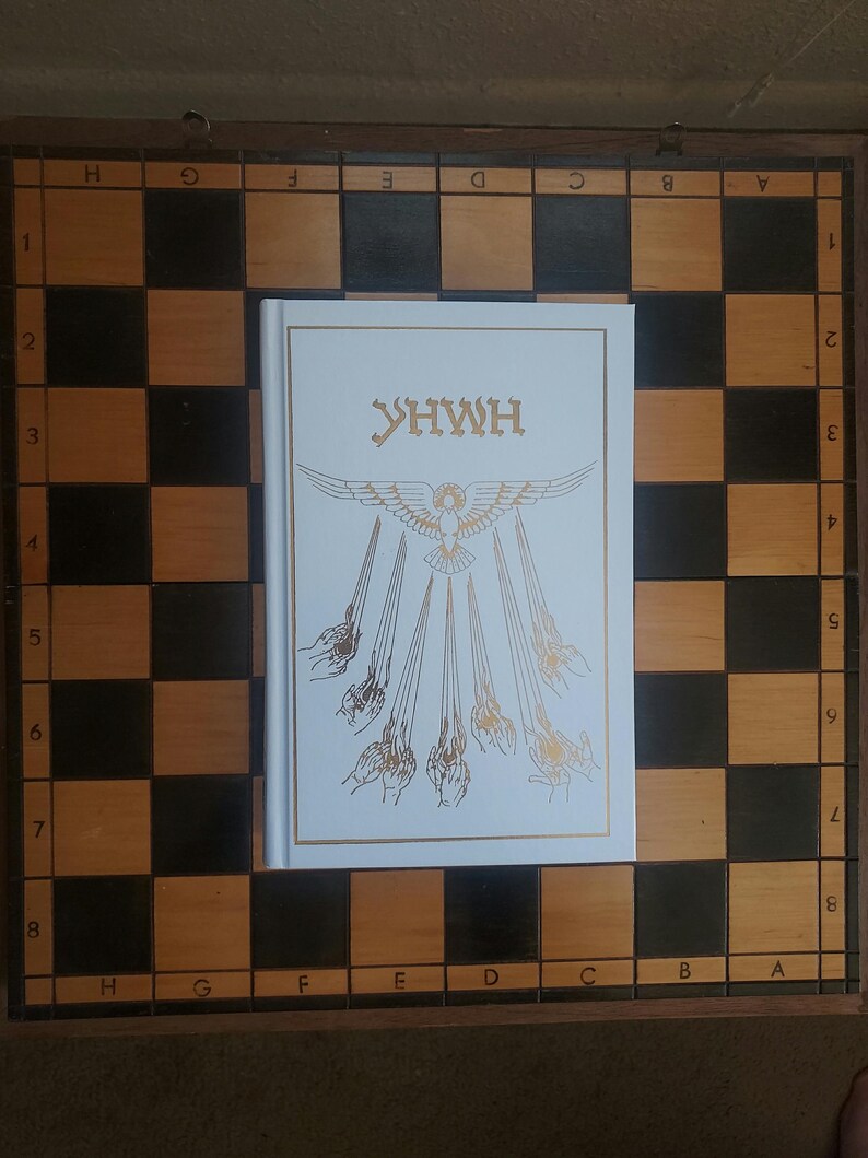 May include: A white book with gold lettering and a detailed illustration of a bird and hands, resting on a wooden chessboard with black and tan squares. The book's title is "YHWH".
