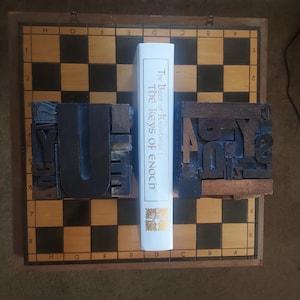 May include: A white book titled "The Book of Knowledge: The Keys of Enoch" stands between two wooden letterpress blocks on a chessboard. The blocks are dark brown with visible letterforms. The chessboard has a brown wooden frame.