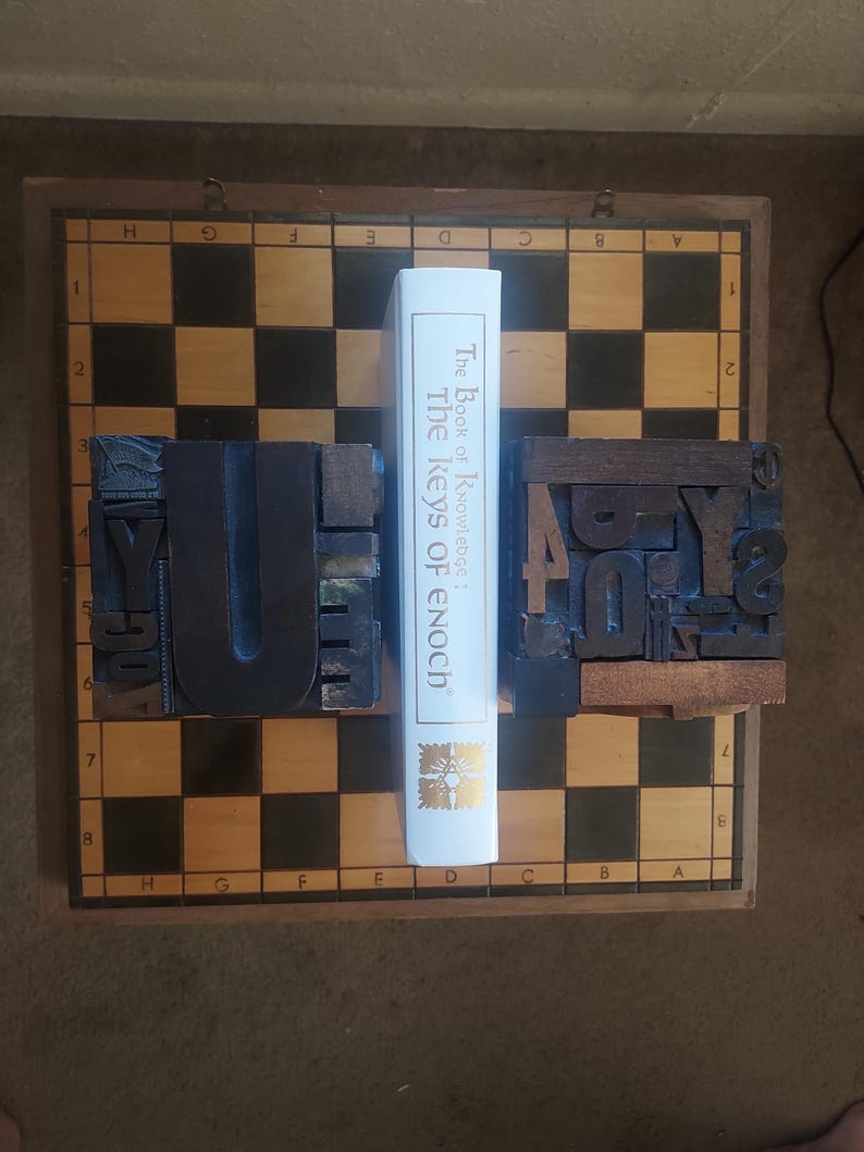 May include: A white book titled "The Book of Knowledge: The Keys of Enoch" stands between two wooden letterpress blocks on a chessboard. The blocks are dark brown with visible letterforms. The chessboard has a brown wooden frame.
