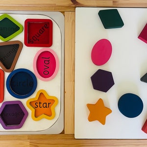 Könnte beinhalten: Ein Holzspielzeug mit verschiedenen Aussparungen in Form von geometrischen Figuren. Die Formen umfassen einen Kreis, ein Quadrat, ein Dreieck, eine Raute, ein Oval, ein Rechteck, ein Sechseck und einen Stern. Die Formen sind mit ihren Namen beschriftet.