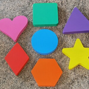 Pode incluir: Sete formas de espuma coloridas: um coração, um quadrado, um triângulo, um círculo, um diamante, um hexágono e uma estrela. As formas são todas de cores diferentes e estão dispostas em círculo sobre uma superfície cinza.
