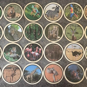 May include: A set of 24 circular coasters displaying images of diverse wildlife, such as a lion, zebra, and panda. Each coaster has a wooden edge and a detailed animal illustration. The coasters are arranged on a grey surface.