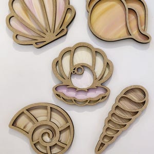 May include: Five wooden seashell cutouts with stained glass inserts. The shells include a scallop, conch, and nautilus design. The stained glass features shades of pink, yellow, and white. The cutouts are arranged on a white surface.