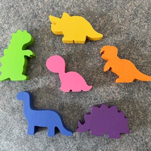 Puede incluir: Seis formas de dinosaurio de madera en diferentes colores: verde, amarillo, rosa, naranja, azul y morado. Los dinosaurios están todos mirando en la misma dirección y están dispuestos en un círculo.