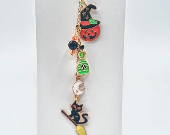May include: A Halloween-themed charm featuring a pumpkin wearing a witch's hat, a candy bag, a crescent moon, and a black cat on a broom. The charm is made of colorful enamel and gold-tone metal. The text "Polishing Gems Handmade" is visible.