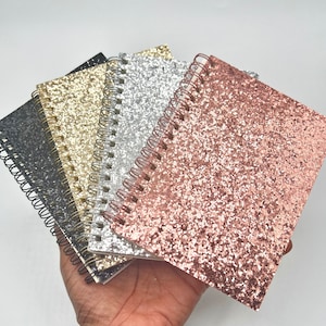 May include: A set of five spiral bound notebooks with glitter covers. The covers are black, gold, silver, and rose gold.
