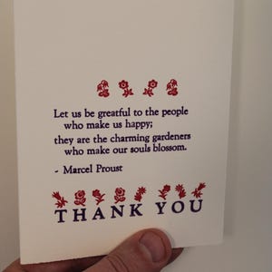 Puede incluir: Una tarjeta blanca con una cita de Marcel Proust impresa en tinta morada. La cita dice: "Seamos agradecidos con las personas que nos hacen felices; son los encantadores jardineros que hacen florecer nuestras almas." La tarjeta también tiene un diseño floral rojo y las palabras "GRACIAS" impresas en tinta morada.