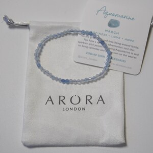 May include: A light blue aquamarine gemstone bracelet with a white pouch and a card that reads "Aquamarine March Happiness Love Hope Zodiac Sign Pisces/Aries @arora_london rorlondon.com".