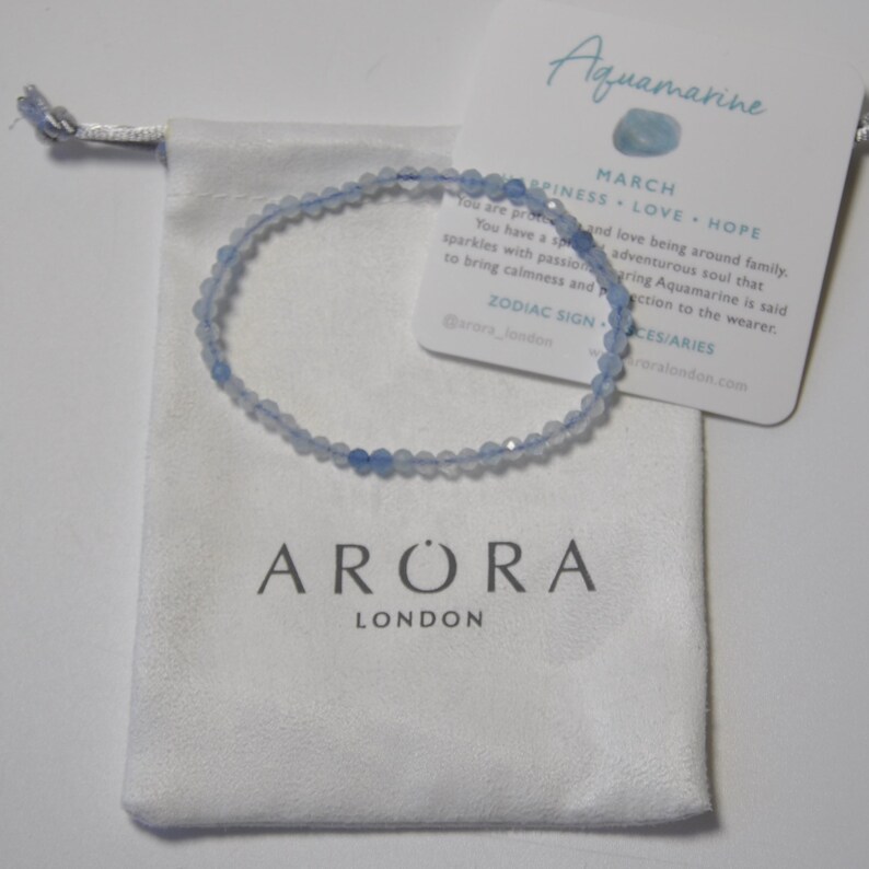 May include: A light blue aquamarine gemstone bracelet with a white pouch and a card that reads "Aquamarine March Happiness Love Hope Zodiac Sign Pisces/Aries @arora_london rorlondon.com".