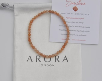 May include: A beaded sunstone bracelet with a drawstring pouch and a card. The bracelet is made of small, round, peach-colored beads. The card reads "Sunstone" and lists the crystal's properties. The pouch is white with the brand name "ARORA LONDON".