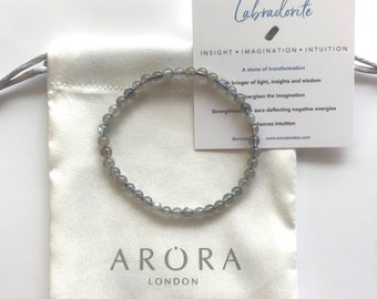May include: A gray labradorite gemstone bracelet on a white fabric pouch with the text "ARORA LONDON" printed on the pouch. The card in the background reads "Labradorite", "INSIGHT IMAGINATION INTUITION", "A stone of transformation", "A bringer of light, insights and wisdom", "Energises the imagination", "Strengthens aura deflecting negative energies", "Enhances intuition", "@aroralondon", "www.aroralondon.com".