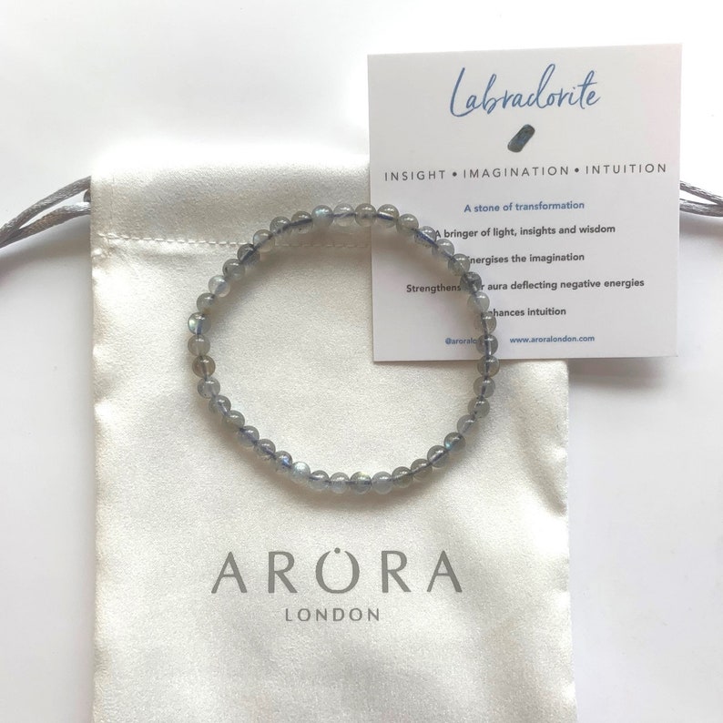 May include: A grey labradorite gemstone bracelet on a white fabric pouch with the text "ARORA LONDON" printed on the pouch. The card in the background reads "Labradorite", "INSIGHT IMAGINATION INTUITION", "A stone of transformation", "A bringer of light, insights and wisdom", "Energises the imagination", "Strengthens aura deflecting negative energies", "Enhances intuition", "@aroralondon", "www.aroralondon.com".