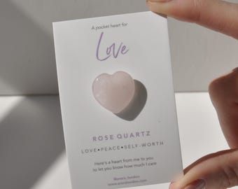 Op de afbeelding: Een roze rozenkwarts hartvormig kristal op een witte kaart met de tekst "A pocket heart for Love" en "ROSE QUARTZ LOVE PEACE SELF-WORTH" en "Here's a heart from me to you to let you know how much I care @arura_london www.aroralandan.com"