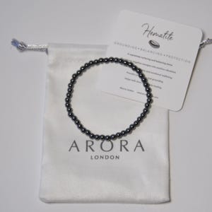 Może przedstawiać: Czarna bransoletka z koralików hematytu na białym aksamitnym woreczku. Woreczek ma nadrukowany tekst "ARORA LONDON". Na tle widoczna jest karta z tekstem "Hematite GROUNDING - BALANCING - PROTECTION".