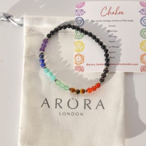 Può includere: Un braccialetto di perline con i colori dei chakra, esposto con un sacchetto bianco con coulisse e una carta che elenca i sette centri energetici del corpo. Il braccialetto presenta perline di vari colori, tra cui viola, blu, verde e rosso. La borsa ha il testo "ARORA LONDON".