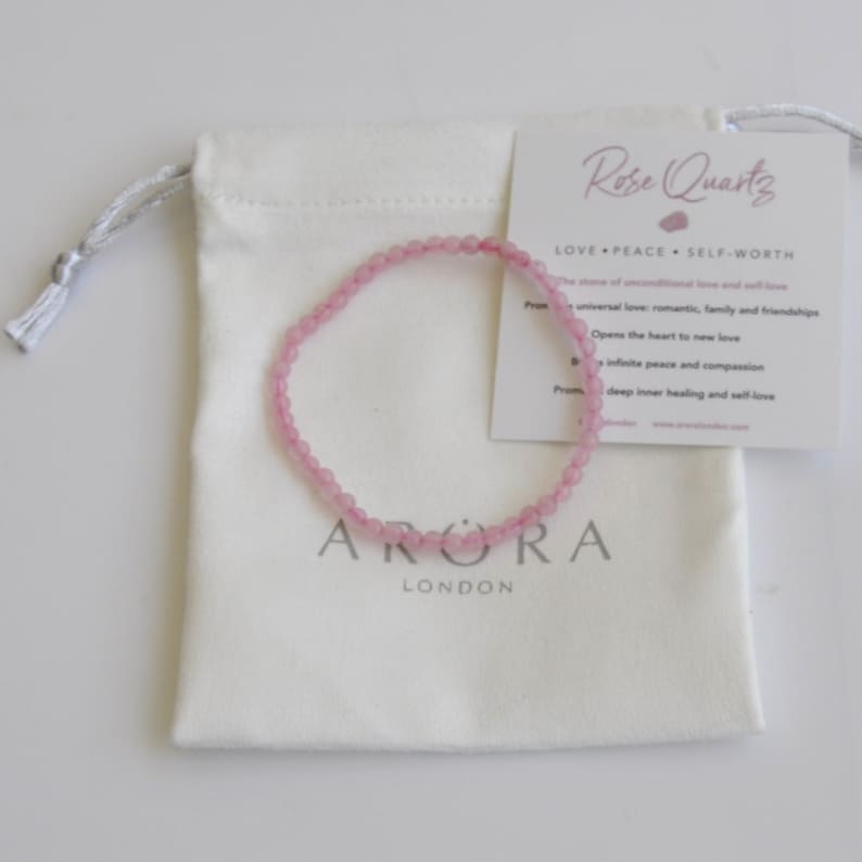 Op de afbeelding: Een roze rozenkwarts armband met een witte trekkoordtas. De tas heeft de tekst "ARORA LONDON" erop gedrukt. De kaart die aan de tas is bevestigd, luidt "Rose Quartz LOVE PEACE SELF-WORTH The stone of unconditional love and self-love Prom universal love: romantic, family and friendships Opens the heart to new love A infinite peace and compassion Prom deep inner healing and self-love"