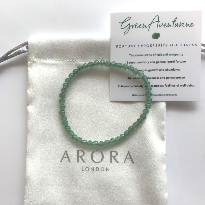 Pu&ograve; includere: Un delicato braccialetto di avventurina verde con piccole perline rotonde &egrave; appoggiato su un sacchetto di raso bianco. &Egrave; visibile una carta con il testo "Green Aventurine" e informazioni sul prodotto. Il braccialetto &egrave; una pietra classica di fortuna e prosperit&agrave;.