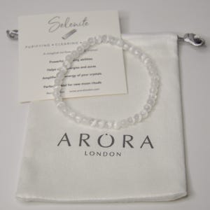 Puede incluir: Un brazalete de cristal de selenita blanco y una bolsa con cordón. La bolsa tiene el texto "ARORA LONDON" impreso. También hay una tarjeta con el texto "Selenerg" y "PURIFYING CLEARING".