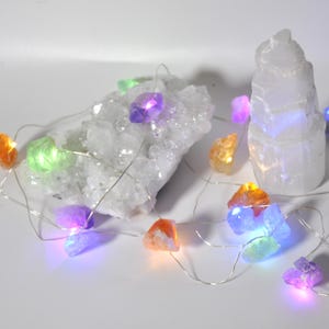 May include: A decorative string of fairy lights featuring crystal-shaped bulbs in various colours. The lights are placed near a large, white crystal cluster and a layered crystal formation. The bulbs emit shades of purple, orange, green, and blue.