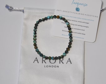 May include: Turquoise gemstone beaded bracelet with brown and black accents. The bracelet is in a white drawstring bag with the word "ARORA LONDON" printed on it. The card next to the bag reads "Turquoise ANTI-ANXIETY CALMING SOOTHING".