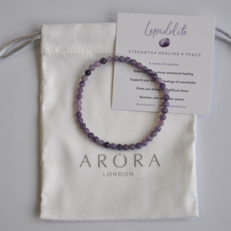 Puede incluir: Un brazalete de cristal de lepidolita morado con las palabras "Lepidolita", "Fuerza", "Sanaci&oacute;n", "Paz", "Una piedra de transici&oacute;n", "Equilibra", "nutre la sanaci&oacute;n emocional", "Te apoya a trav&eacute;s", "sentimientos de abrumamiento", "Te da fuerza", "tiempos dif&iacute;ciles", "Concede calma y paz interior", "ARORA" y "LONDRES" impresas en una tarjeta blanca. El brazalete est&aacute; en una bolsa de cord&oacute;n blanco.