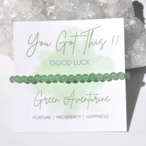 Puede incluir: Una pulsera de cuentas de aventurina verde se muestra en una tarjeta blanca. La tarjeta tiene el texto "You Got This !! Good Luck" y "Green Aventurine". La tarjeta también incluye las palabras "Fortune | Prosperity | Happiness".