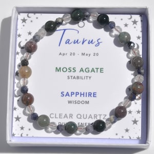 May include: A beaded bracelet featuring a mix of clear quartz, moss agate, and sapphire stones. The bracelet is displayed in a white box with text that reads "Taurus" and "Apr 20 - May 20". The card also lists the properties of the stones.