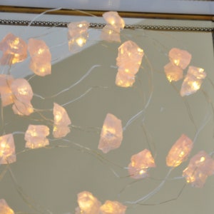 May include: A string of fairy lights featuring pale pink, irregularly shaped crystals. Each crystal is illuminated with a warm, soft glow, strung on delicate silver wire. The crystals are reflected in a mirror.