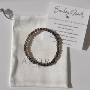 May include: A smoky quartz beaded bracelet, displayed with a white drawstring pouch and a card detailing the stone's properties. The bracelet's beads are a dark brown hue. The card reads "Smokey Quartz".