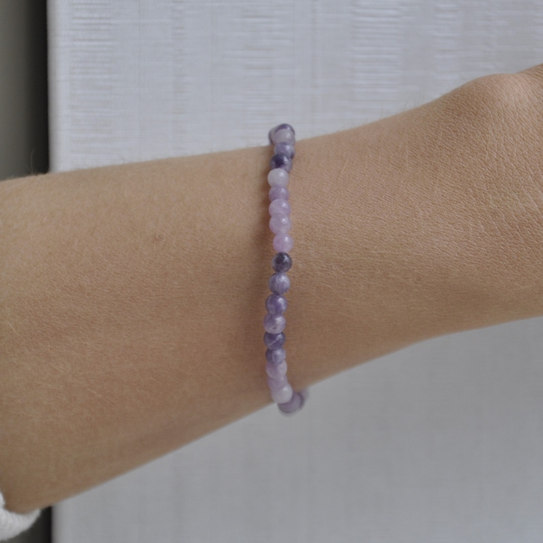 Puede incluir: Un brazalete de cuentas de piedras preciosas de color p&uacute;rpura y lavanda. El brazalete est&aacute; hecho con peque&ntilde;as cuentas redondas que est&aacute;n enhebradas en un cord&oacute;n el&aacute;stico transparente.