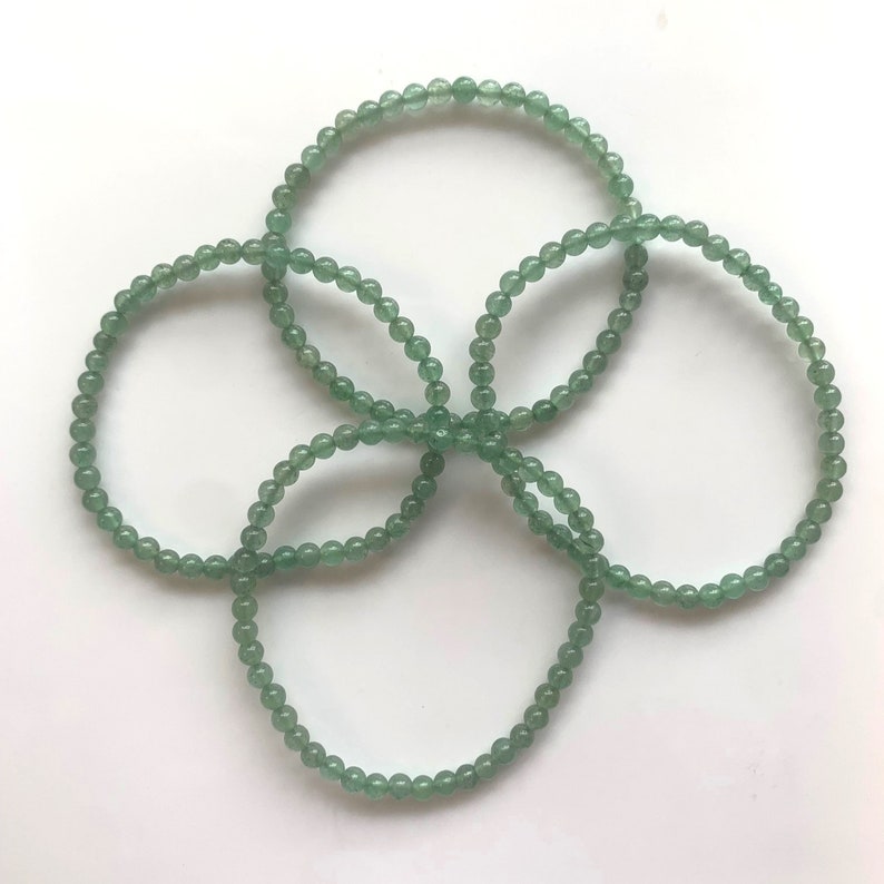 Pu&ograve; includere: Quattro braccialetti di perline verdi disposti a forma di fiore. Ogni braccialetto &egrave; composto da piccole perline rotonde verde chiaro. I braccialetti sono esposti su una superficie bianca, creando una presentazione semplice ma elegante.