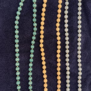 May include: Three long beaded necklaces arranged vertically. The left necklace features green marbled beads, the center necklace has light orange marbled beads, and the right necklace consists of light blue marbled beads, all separated by small gold spacers.